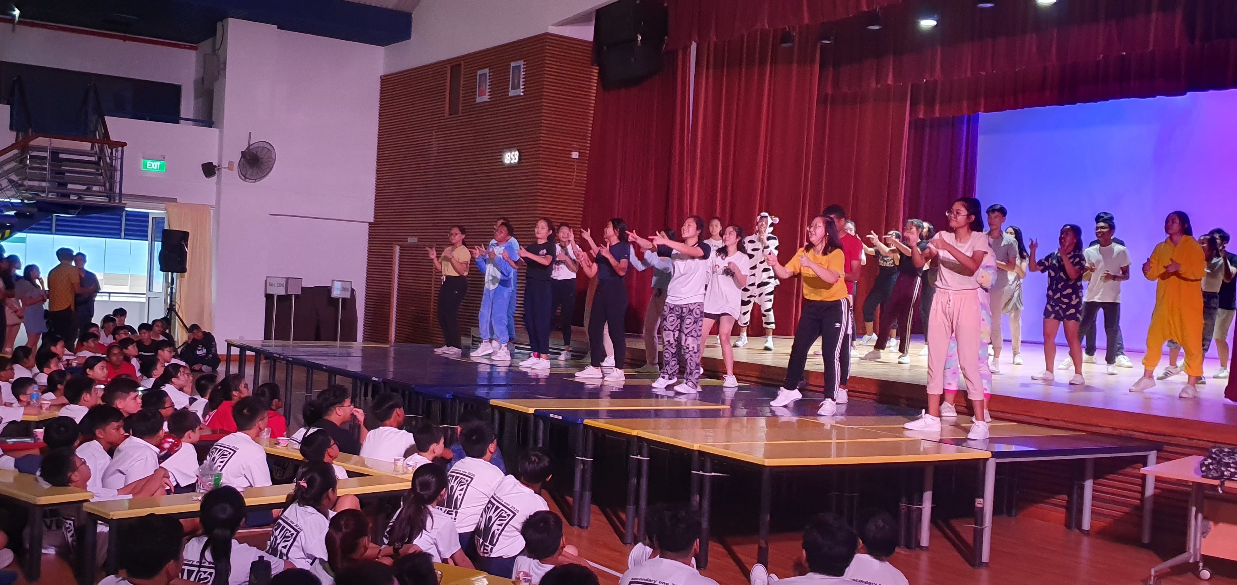Students in costumes perform a dance on a school hall stage while a large seated audience watches.
